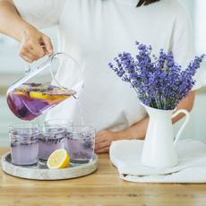 A woman pours lavender tea into five small cups next to a jar of honey and a pitcher of lavender flowers